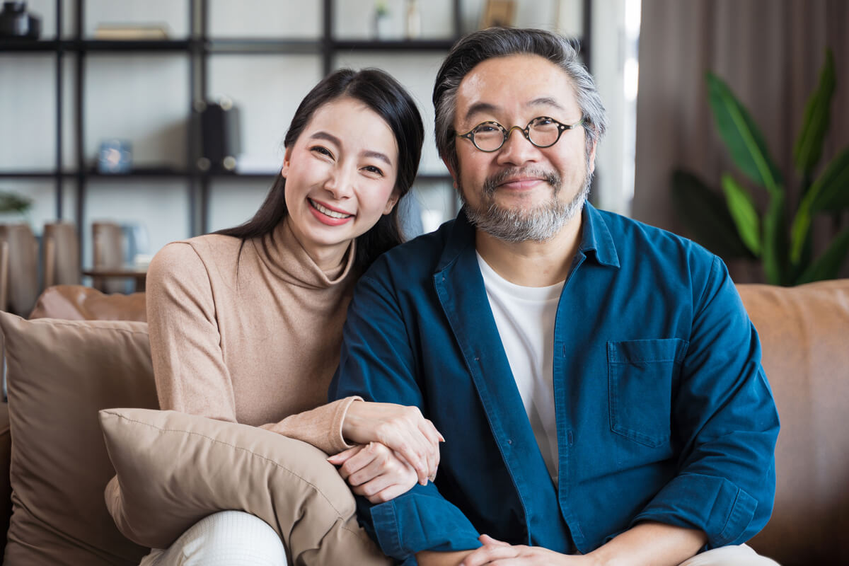 An Asian couple sitting on a couch, smiling and looking at each other affectionately.