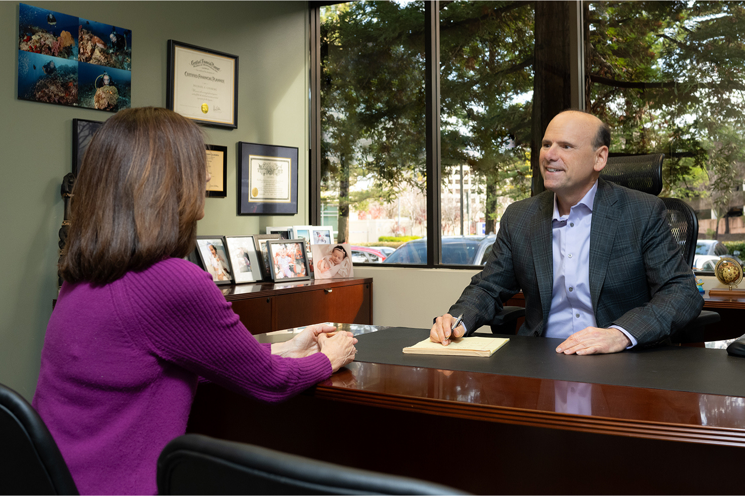 A man in a suit smiles while sitting at an office desk, engaging with a woman in a purple sweater. Certificates and family photos are displayed nearby.