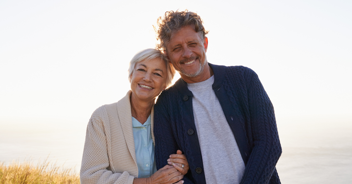 Smiling older couple embracing outdoors, standing on a grassy hill with a serene sea and clear sky in the background. Warm, joyful atmosphere.
